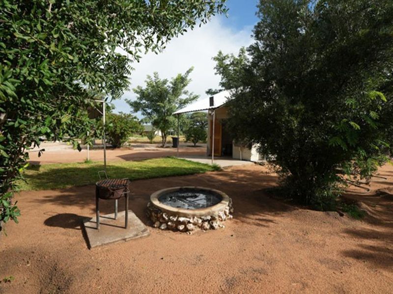 Outdoor garden area featuring a stone-clad fire pit, a concrete patio slab, and a covered outdoor structure set within a natural farm landscape.