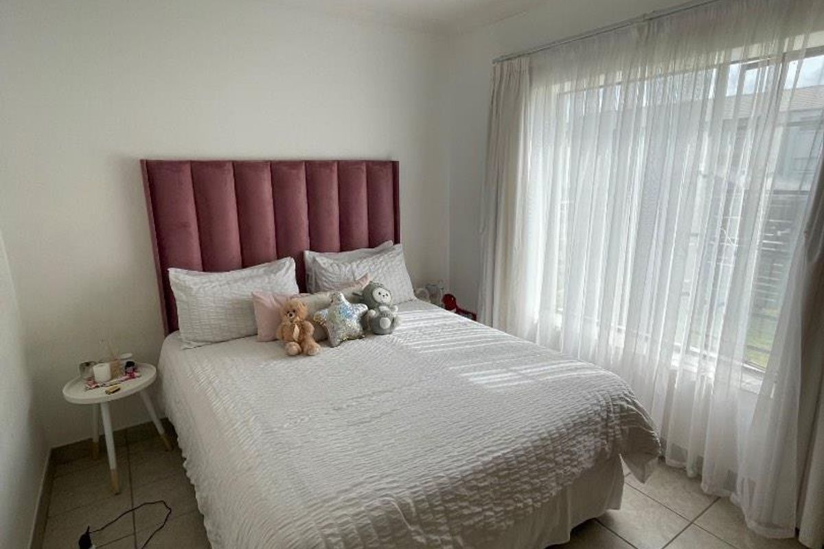 A bright bedroom featuring neutral tiled flooring, a ceiling-mounted spotlight fixture, and large windows dressed with sheer curtains.