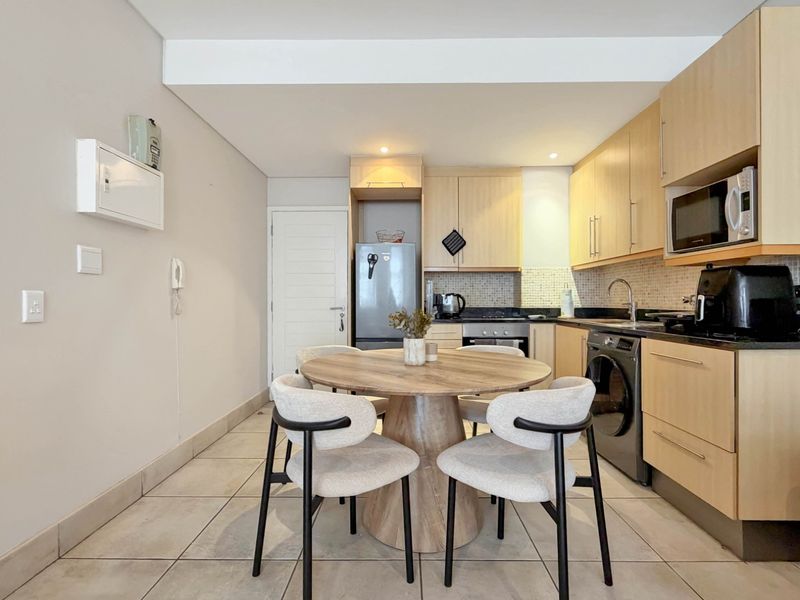Open-plan kitchen featuring light-wood cabinetry, granite countertops, mosaic backsplash, and integrated appliances, seamlessly flowing into a dining area with tiled flooring.