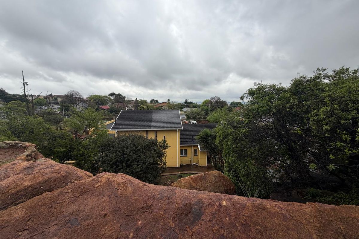 An elevated aerial view of a yellow residential house with a dark tiled roof, surrounded by mature trees and a rocky foreground.