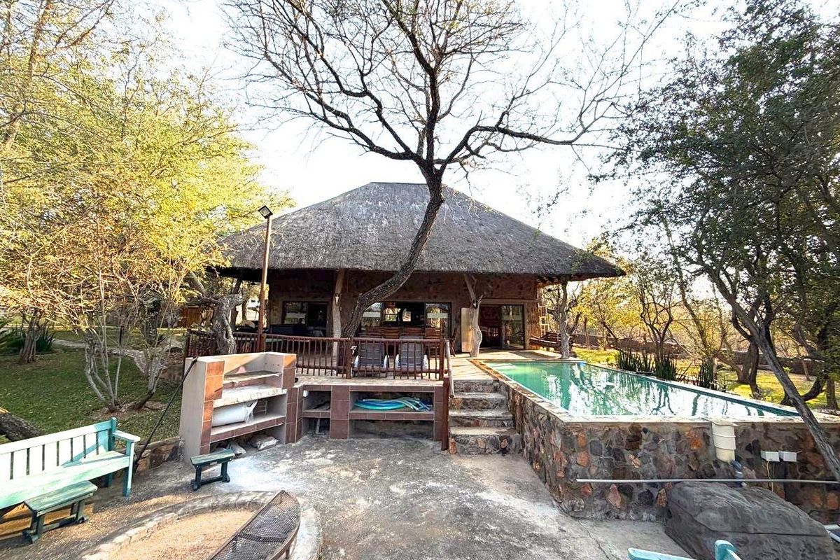 Rear exterior view featuring a thatched-roof dwelling, raised timber deck, swimming pool with stone surround, and a built-in braai area in a natural setting.