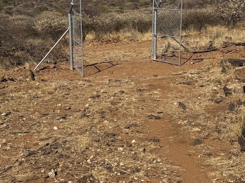 A view of a metal chain-link gate providing access to a vacant agricultural plot with natural landscape and mountain views.