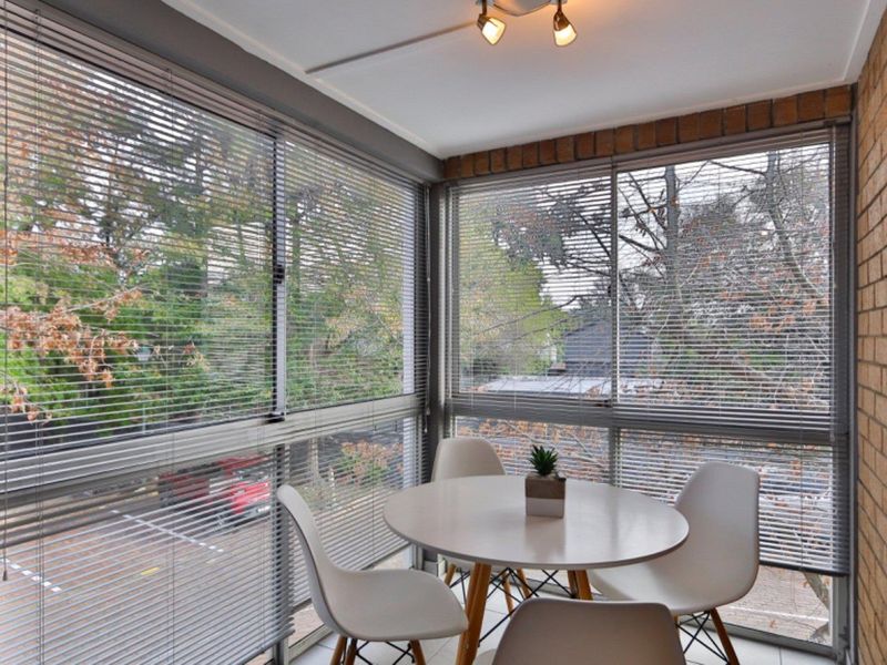 A bright enclosed balcony or sunroom featuring large windows with venetian blinds, exposed brick wall detailing, and modern ceiling track lighting.