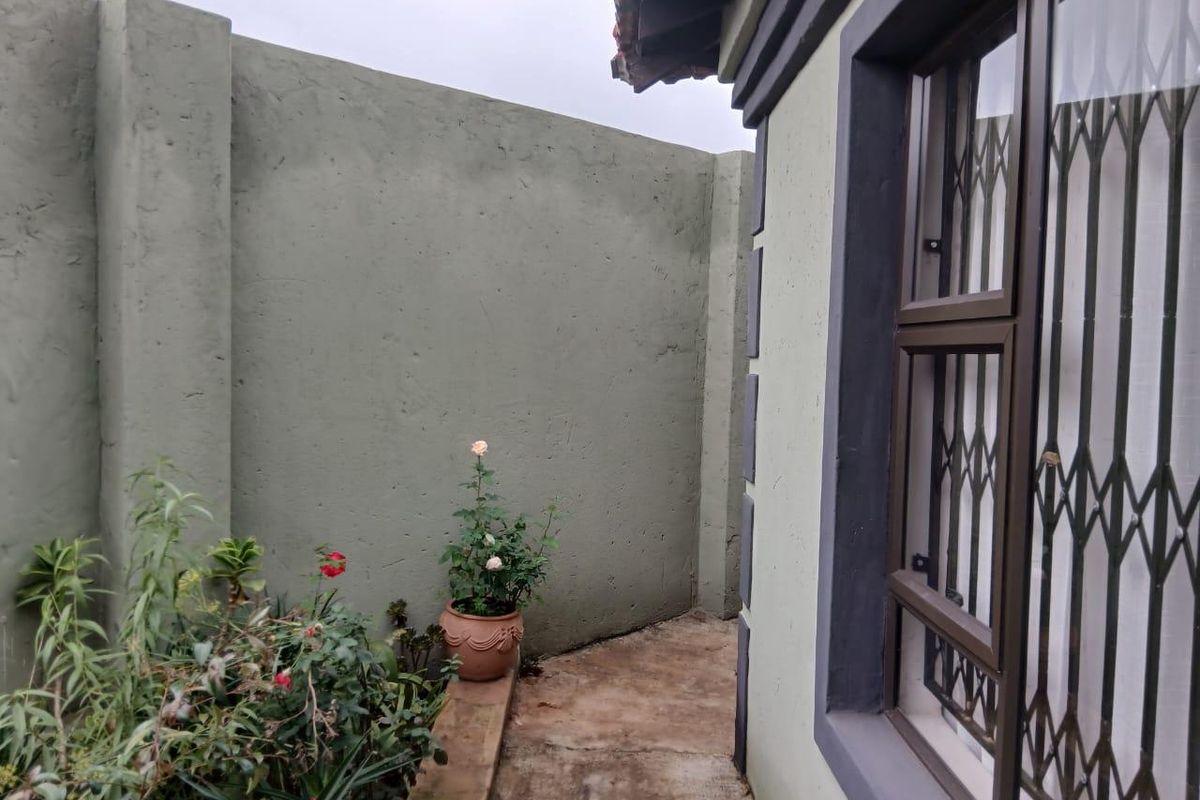 A side exterior view showing a concrete boundary wall, a paved walkway, and a window fitted with security bars.
