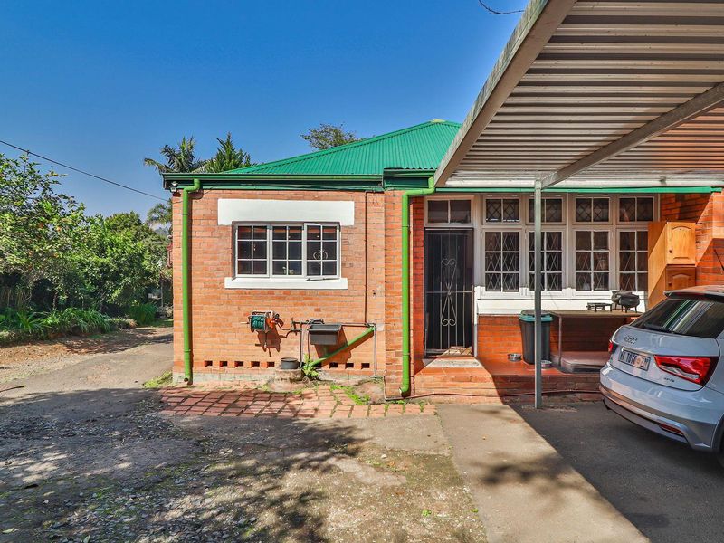 Rear view of a brick house featuring a green corrugated metal roof, a covered carport, and established garden surroundings.