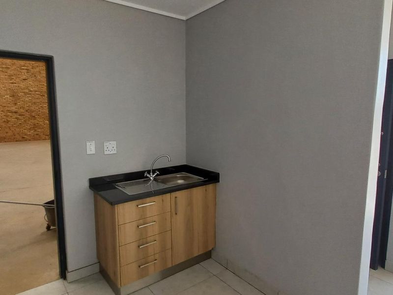 A clean, functional scullery area featuring modern wood-finish cabinetry, a stainless steel sink, and a durable stone countertop, adjacent to a large industrial warehouse space.