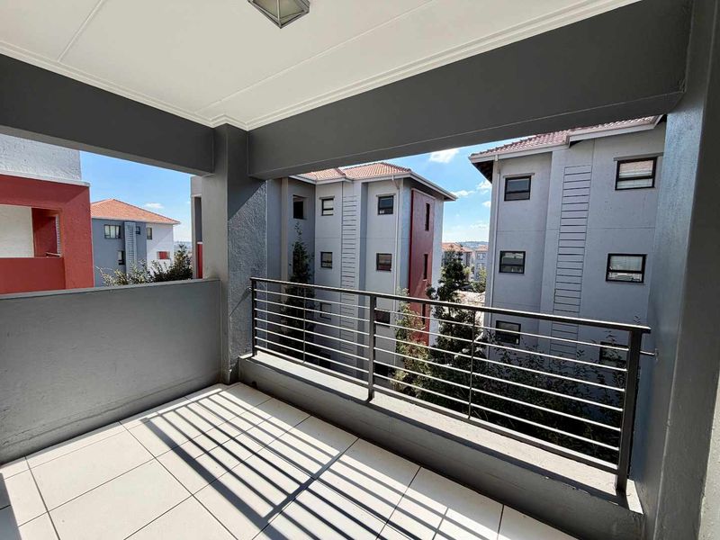 A clean, covered balcony featuring tiled flooring and a metal balustrade, offering an open view of the surrounding apartment complex.