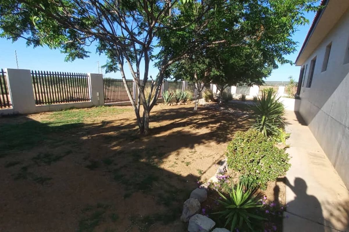 A rear garden area featuring a concrete walkway, perimeter wall with security fencing, and established trees.
