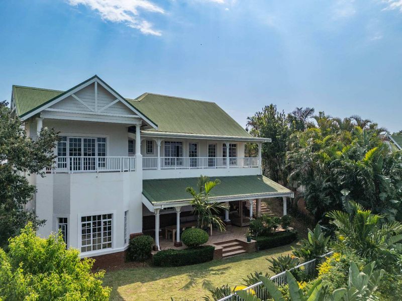 Rear view of a two-story residence featuring a green corrugated metal roof, a spacious covered patio, and an upper-level balcony overlooking a landscaped garden.