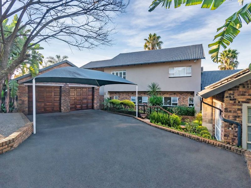 Front exterior view of a multi-level house featuring a double garage, paved driveway with a shade cloth carport, and established garden landscaping.