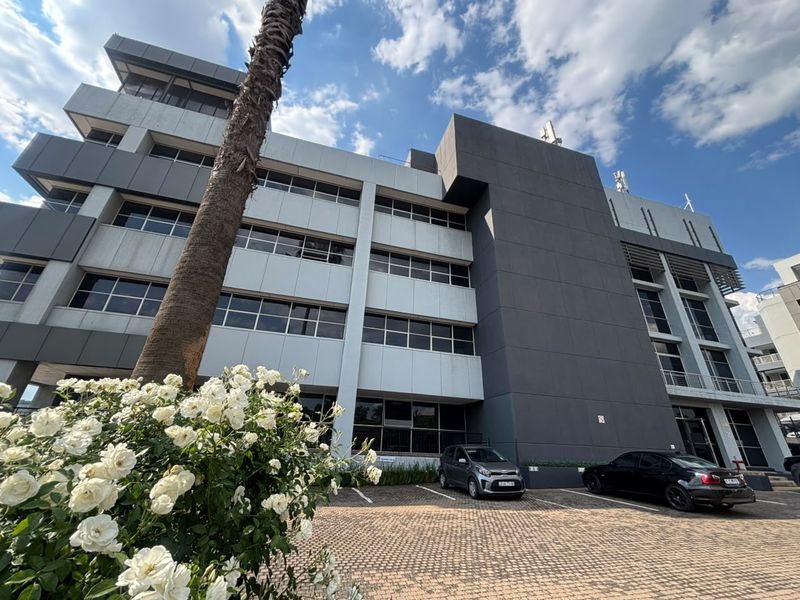 A modern multi-story office building featuring a contemporary facade with aluminum cladding and large windows, situated above a paved parking area.