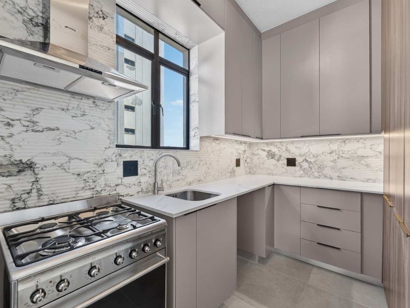 Modern kitchen featuring sleek grey cabinetry, marble-look backsplash, stone countertops, and a stainless steel gas stove with an integrated extractor fan.