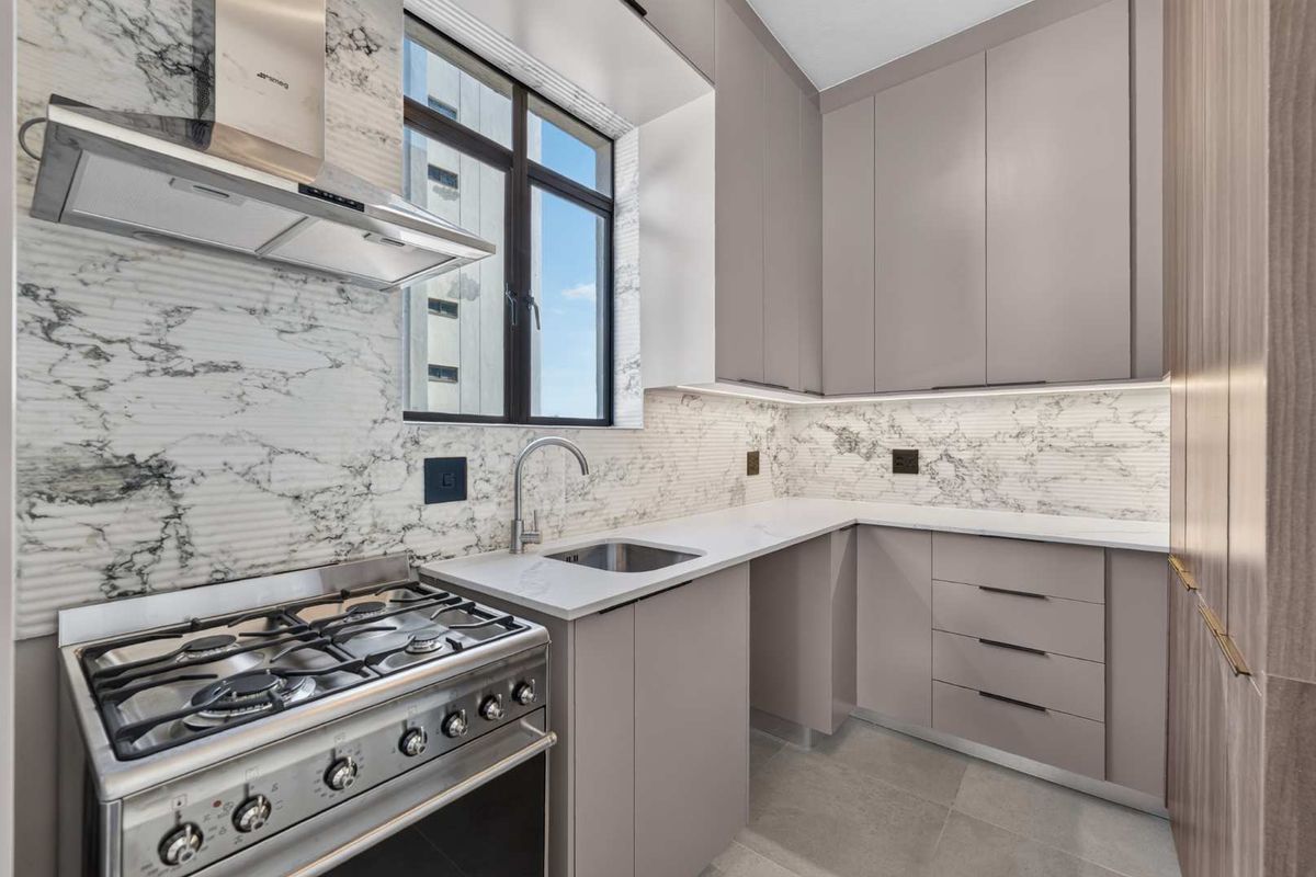 Modern kitchen featuring sleek grey cabinetry, marble-look backsplash, stone countertops, and a stainless steel gas stove with an integrated extractor fan.