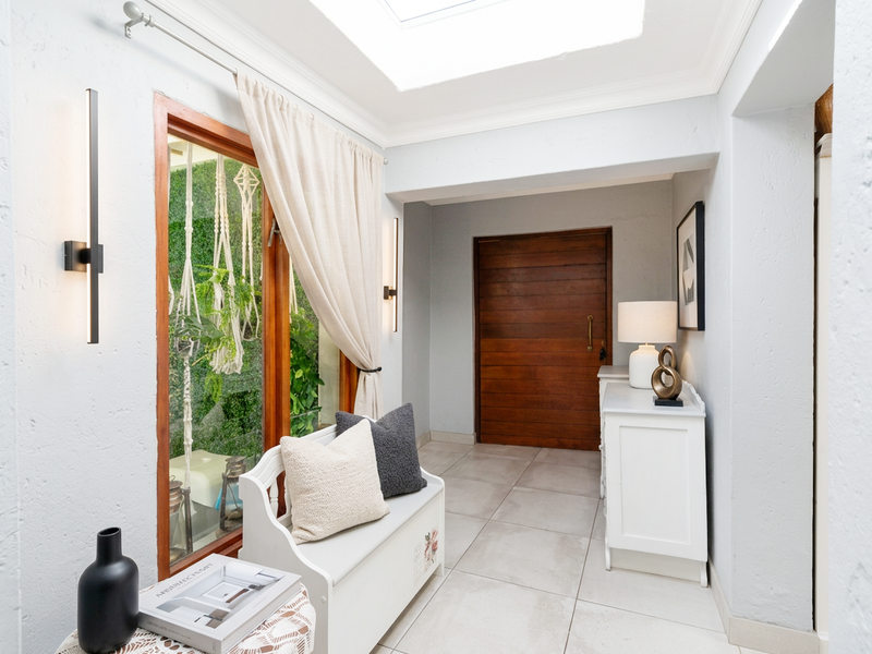 A bright and welcoming entrance hall featuring tiled flooring, a large skylight, and a solid wooden front door with modern wall lighting.