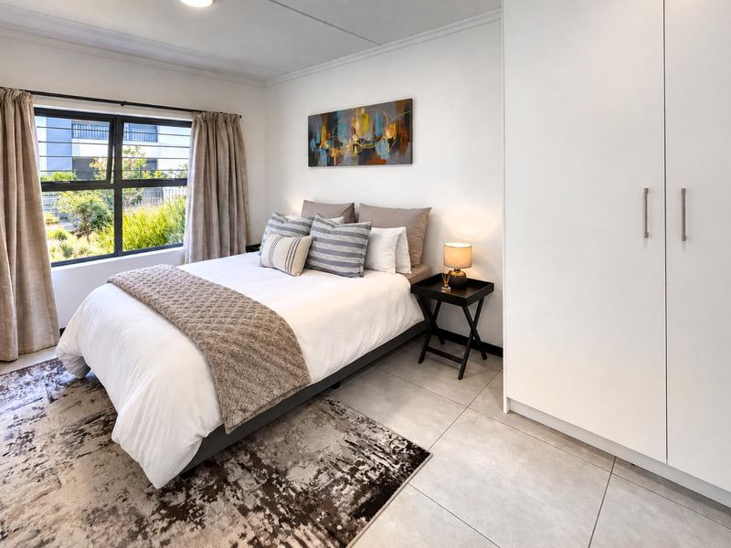 A well-maintained bedroom featuring neutral tiled flooring, a built-in wardrobe, and a window with security bars overlooking the exterior.