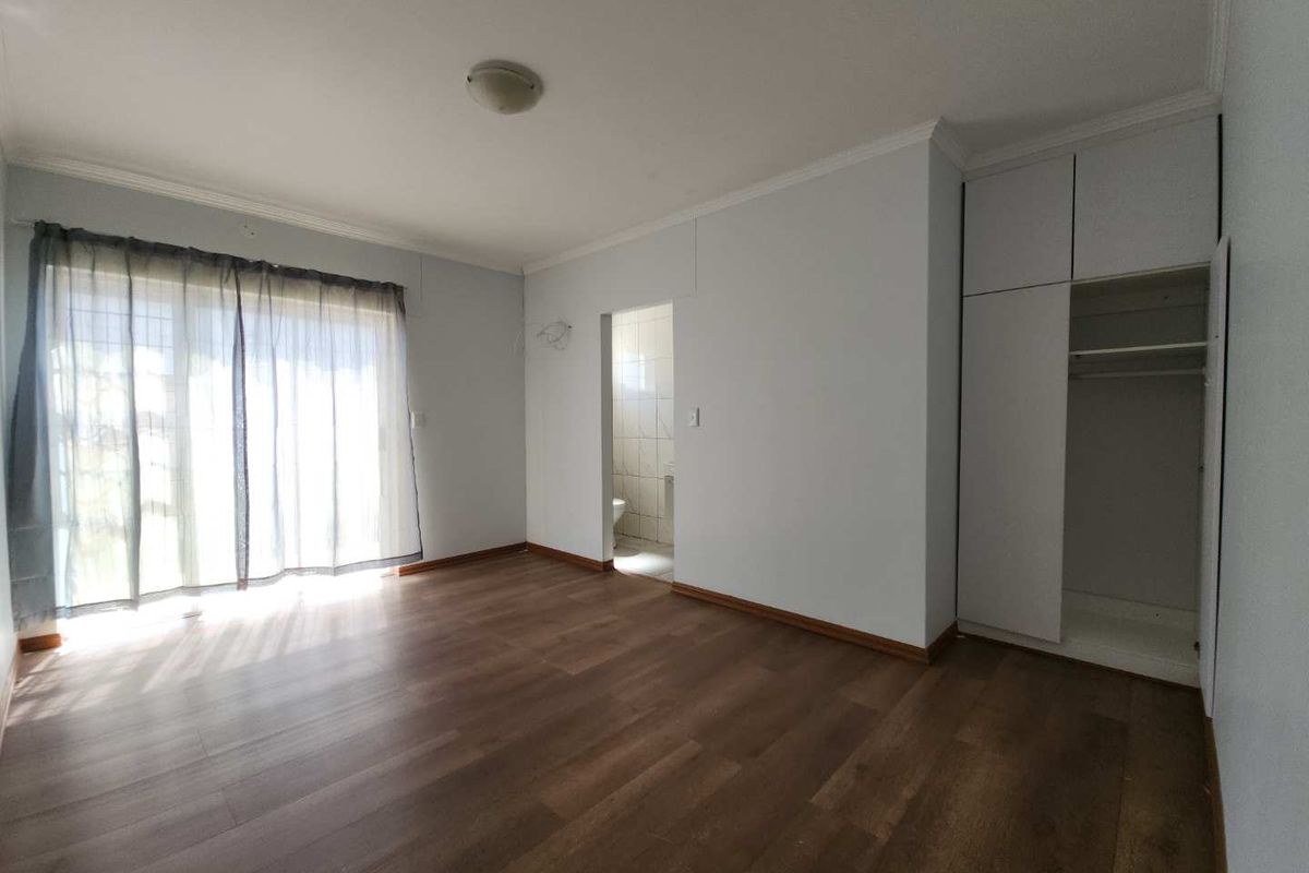 A bright bedroom featuring wood-look flooring, built-in cupboards, and an en-suite bathroom entrance.