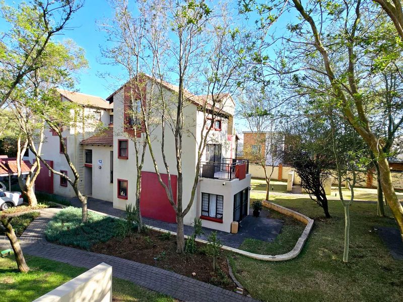 An elevated view of the apartment complex exterior, featuring a paved walkway, landscaped garden areas, and a balcony with security bars.