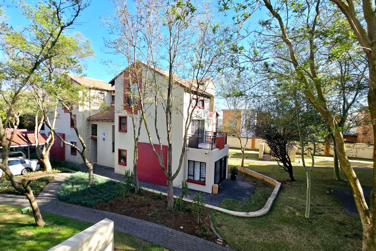 An elevated view of the apartment complex exterior, featuring a paved walkway, landscaped garden areas, and a balcony with security bars.