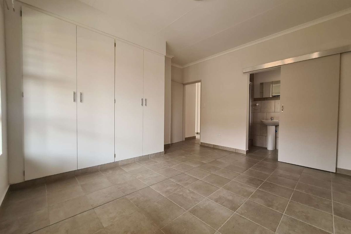 A spacious, empty bedroom featuring neutral tiled flooring, ample built-in cupboard storage, and direct access to an en-suite bathroom via a sliding door.