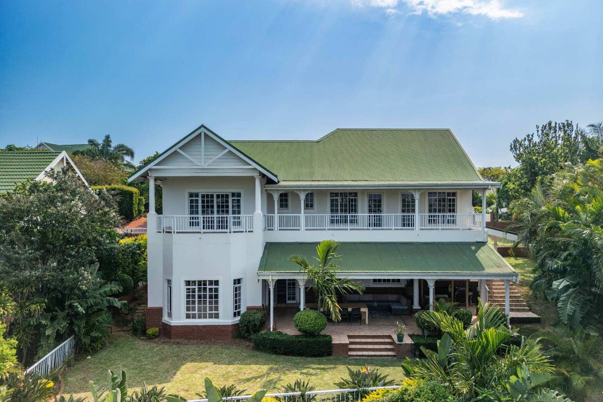 Rear view of a two-story house featuring a green corrugated metal roof, a spacious covered patio, and an upper-level balcony overlooking the garden.