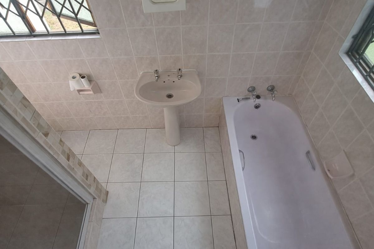 A functional bathroom featuring a pedestal basin, a standard bathtub, and tiled walls and flooring, with security bars on the windows.