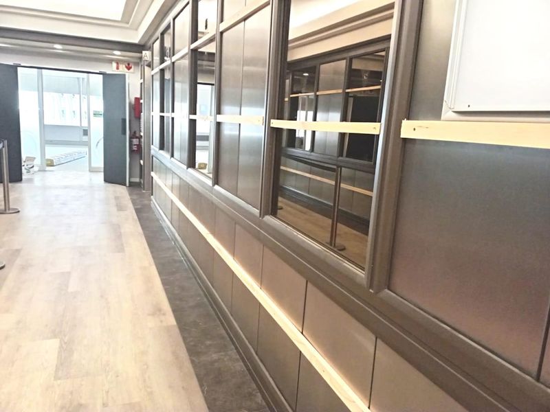 A modern office hallway featuring glass partition walls, wood-look laminate flooring, and recessed ceiling lighting, currently in a clean, vacant state.