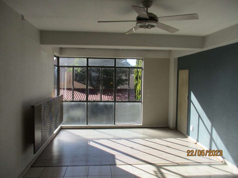 An empty living room featuring a ceiling fan, large metal-framed windows providing natural light, and tiled flooring with a blue accent wall.