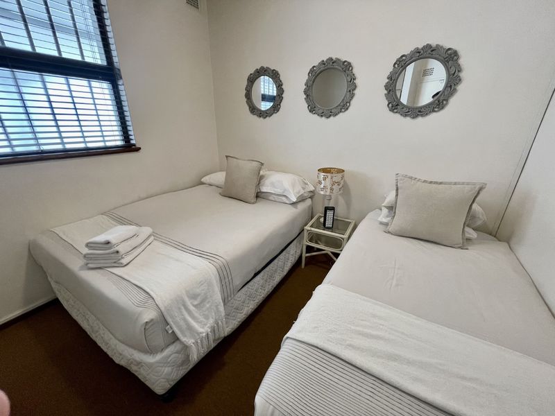 A compact bedroom featuring two single beds, neutral wall finishes, and a window with venetian blinds.