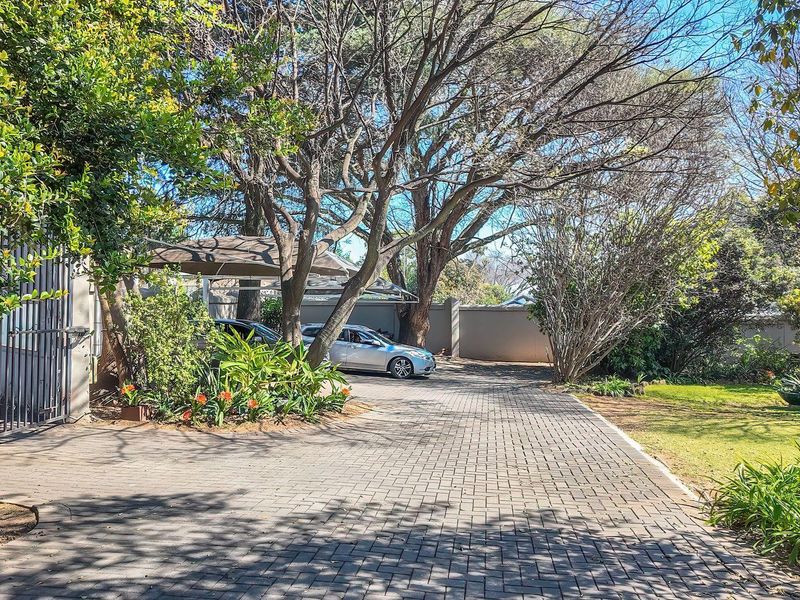 A well-maintained paved driveway leading to a property entrance, featuring mature trees, landscaped garden beds, and a covered carport area.