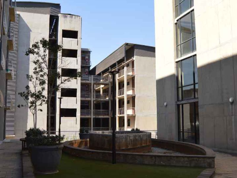 A view of a communal courtyard area between apartment blocks, featuring paved walkways, an artificial lawn, and a central concrete fountain feature.