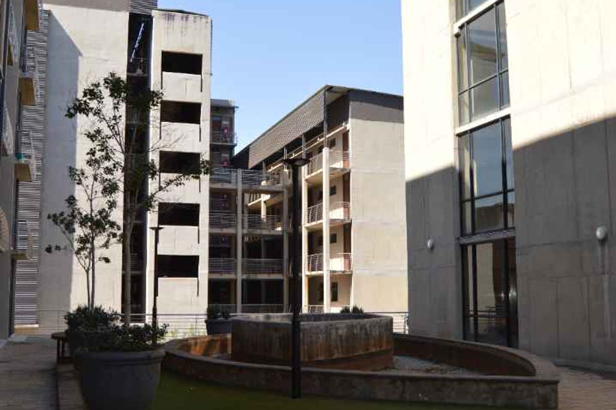 A view of a communal courtyard area between apartment blocks, featuring paved walkways, an artificial lawn, and a central concrete fountain feature.