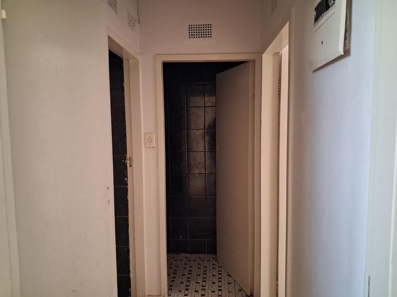 A narrow apartment hallway featuring tiled flooring, an electrical distribution board, and access to adjacent rooms with dark tiled finishes.