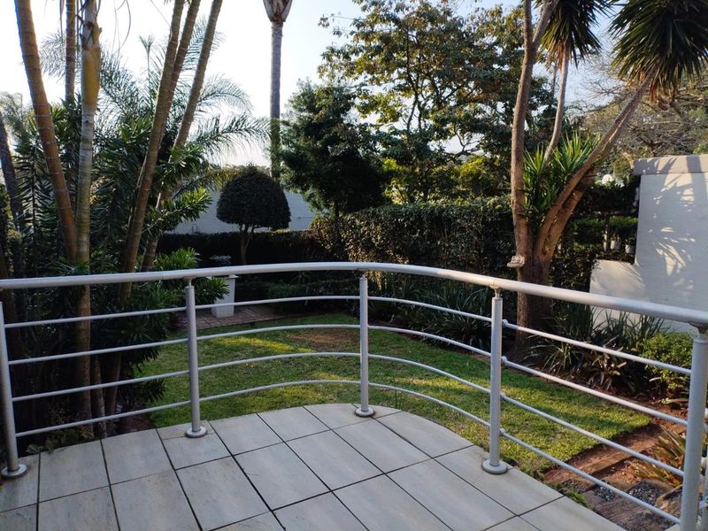 A raised balcony featuring tiled flooring and a metal balustrade, overlooking a lush, established garden area.
