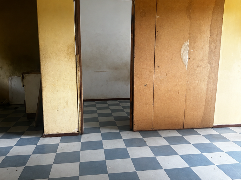 An empty living area featuring checkered vinyl flooring and a wooden sliding door, requiring significant cosmetic renovation and wall repairs.