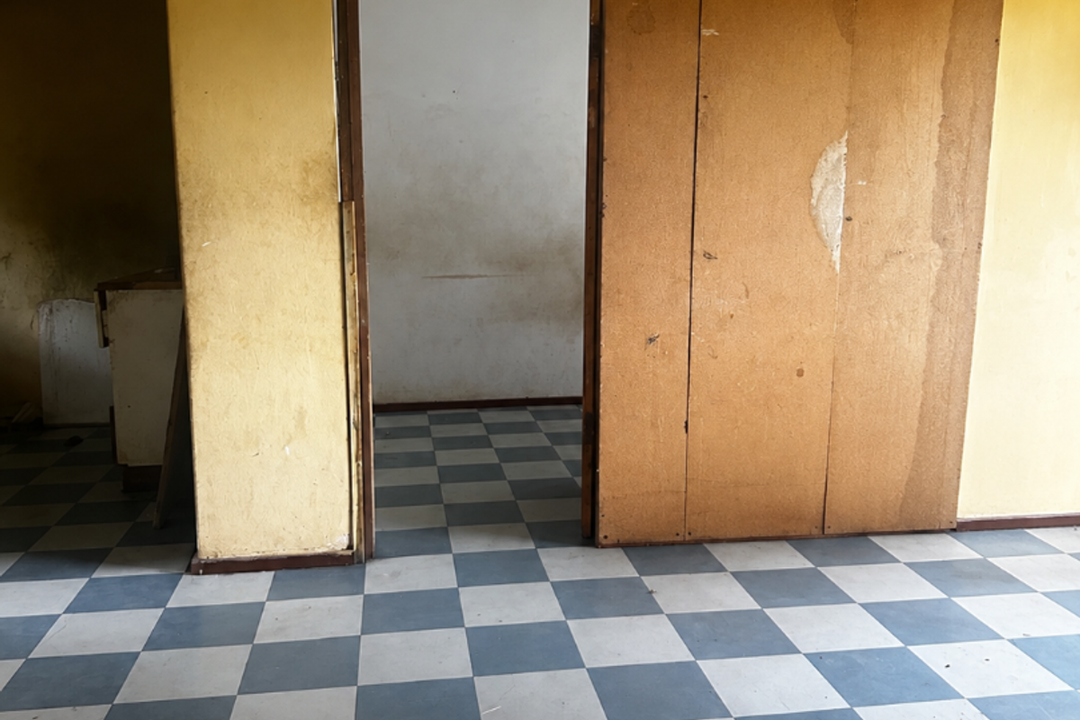 An empty living area featuring checkered vinyl flooring and a wooden sliding door, requiring significant cosmetic renovation and wall repairs.