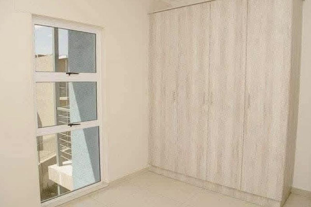 A bright, empty bedroom featuring neutral tiled flooring, a large built-in wardrobe with light wood-grain finish, and a modern aluminium-framed window.