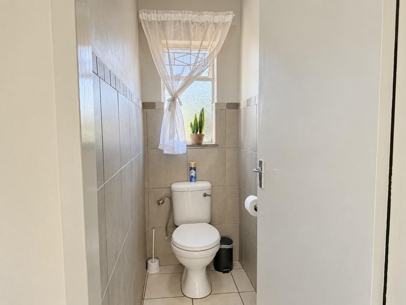 A compact, well-maintained guest toilet featuring neutral ceramic wall and floor tiles and a window with security bars.