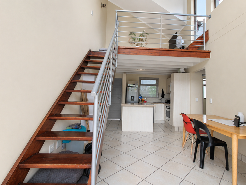 Open-plan living area featuring a timber staircase leading to a mezzanine level, tiled flooring, and a functional kitchen with built-in cabinetry.