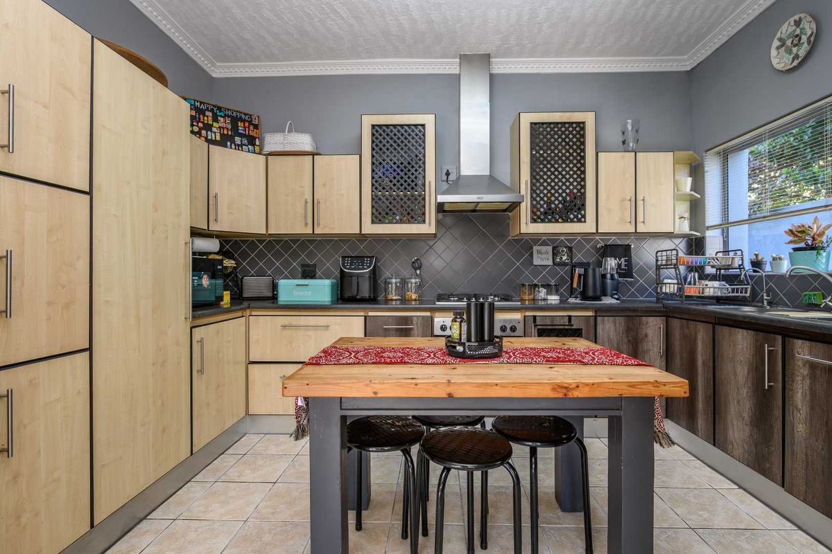 A functional kitchen featuring light wood-tone cabinetry, a stainless steel extractor fan, tiled backsplash, and tiled flooring with a central dining area.