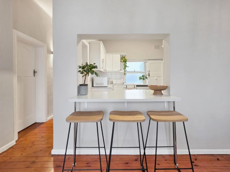 A bright kitchen featuring white cabinetry, a tiled backsplash, and a convenient breakfast bar opening into the living area with hardwood flooring.