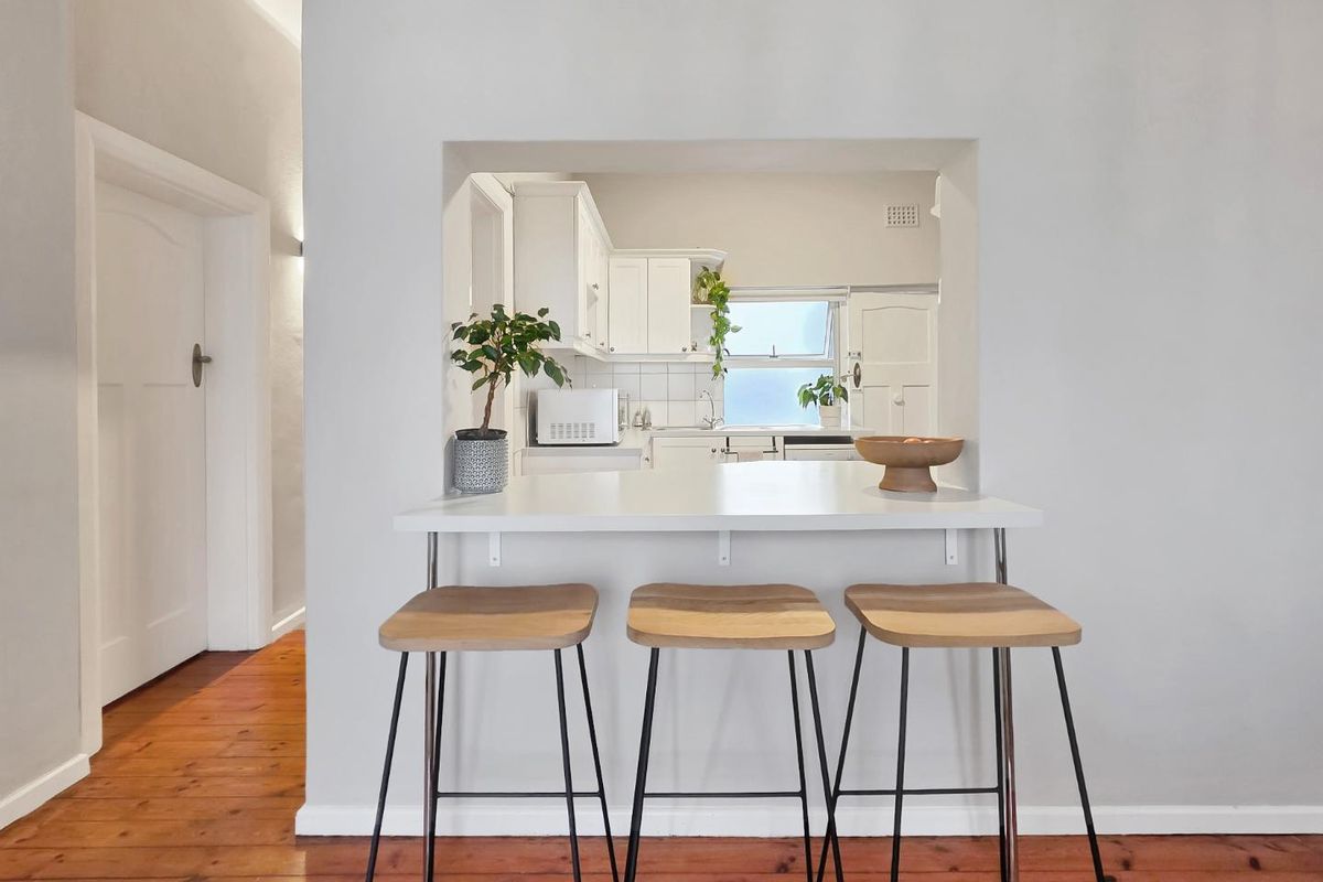 A bright kitchen featuring white cabinetry, a tiled backsplash, and a convenient breakfast bar opening into the living area with hardwood flooring.