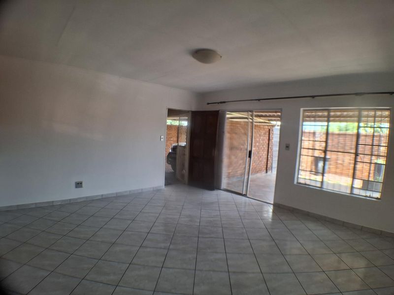 An empty living room featuring neutral ceramic floor tiles, a sliding glass door leading to an outdoor area, and large windows with security bars.