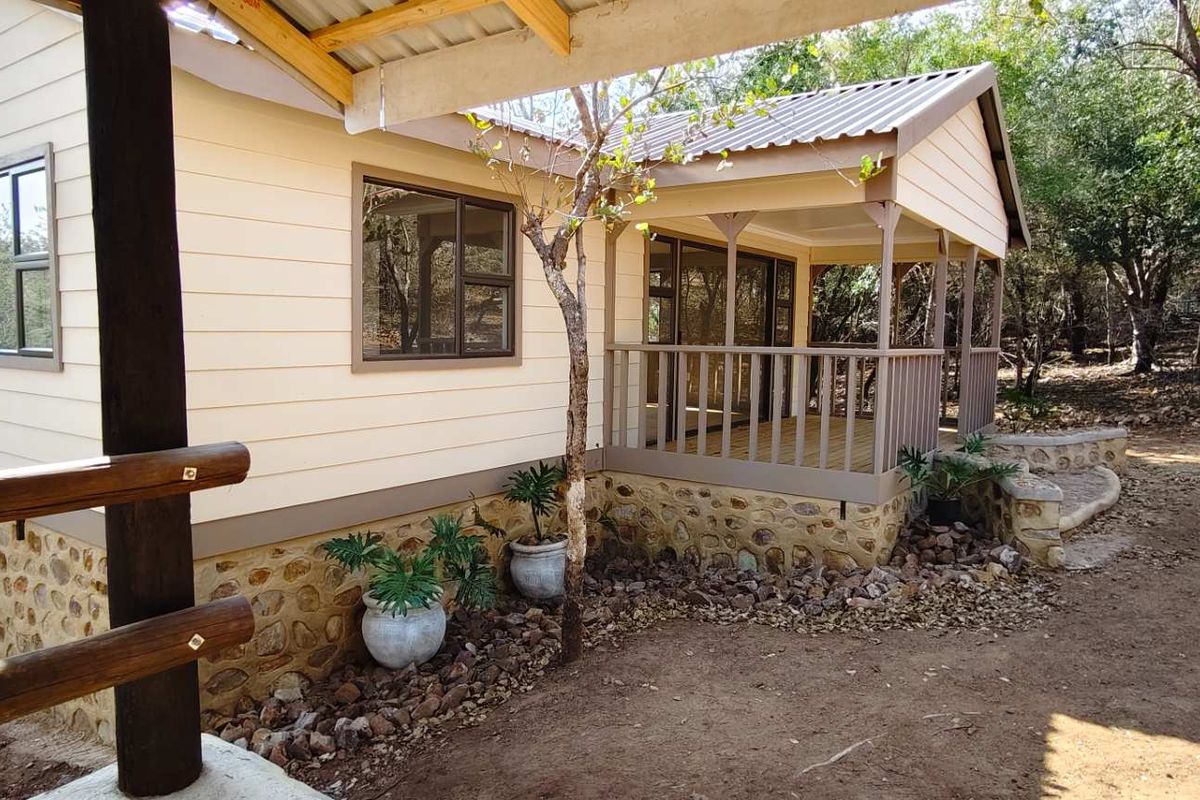 A charming exterior view of a cabin-style home featuring a covered wooden deck, stone foundation detailing, and light-colored siding nestled in a natural setting.
