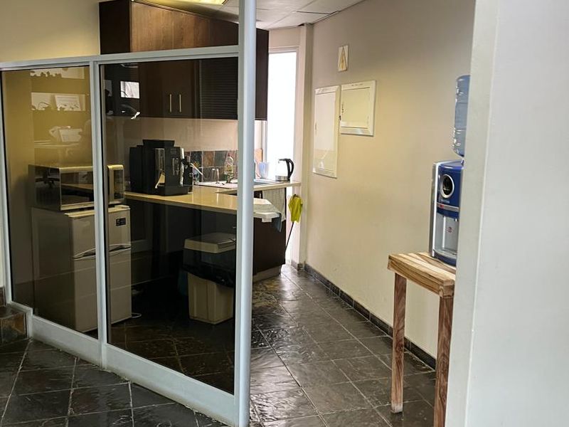 A compact office kitchenette featuring built-in cabinetry, a sink, and tiled flooring, separated from the main office area by a glass partition.