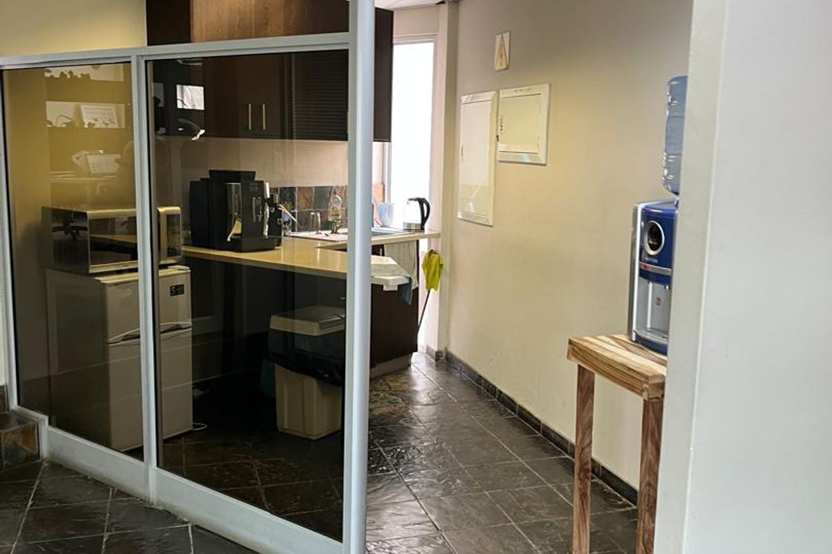 A compact office kitchenette featuring built-in cabinetry, a sink, and tiled flooring, separated from the main office area by a glass partition.