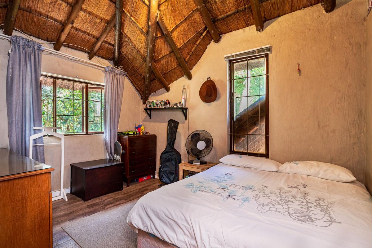 A rustic bedroom featuring a high thatched roof with exposed timber beams, textured walls, and large windows providing natural light.