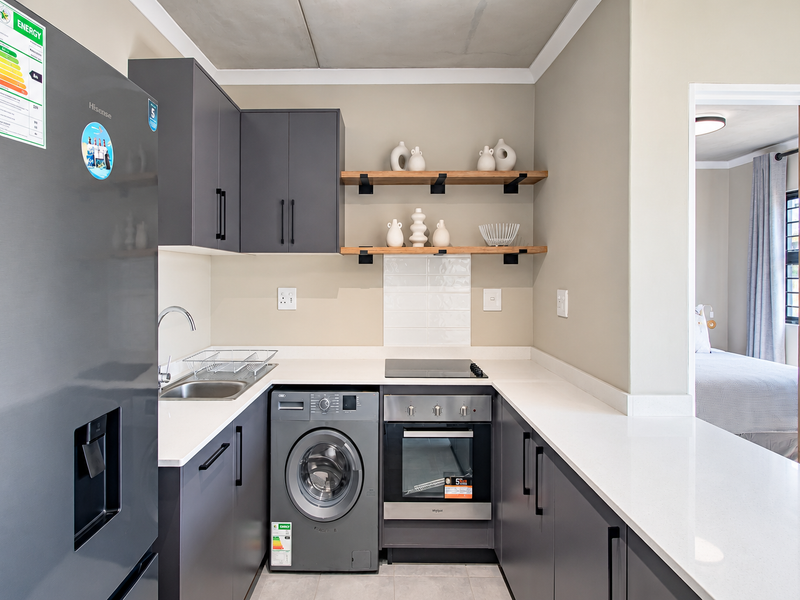 Modern kitchen featuring sleek grey cabinetry, quartz countertops, built-in oven, electric hob, and integrated washing machine, complemented by stylish floating timber shelving.