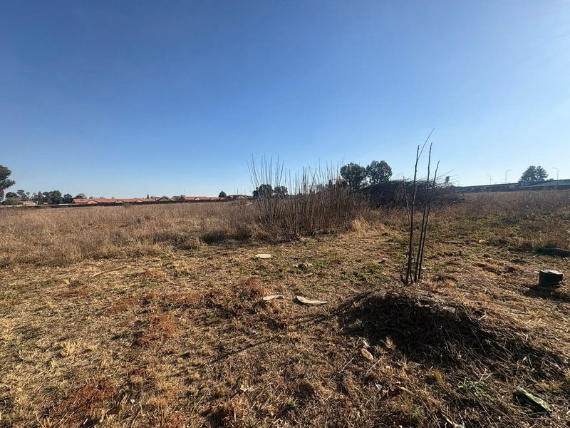 A wide-angle view of a vacant residential plot featuring dry grass and natural vegetation under a clear blue sky.