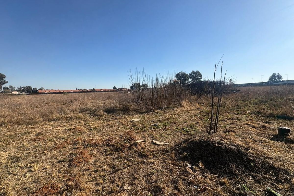 A wide-angle view of a vacant residential plot featuring dry grass and natural vegetation under a clear blue sky.