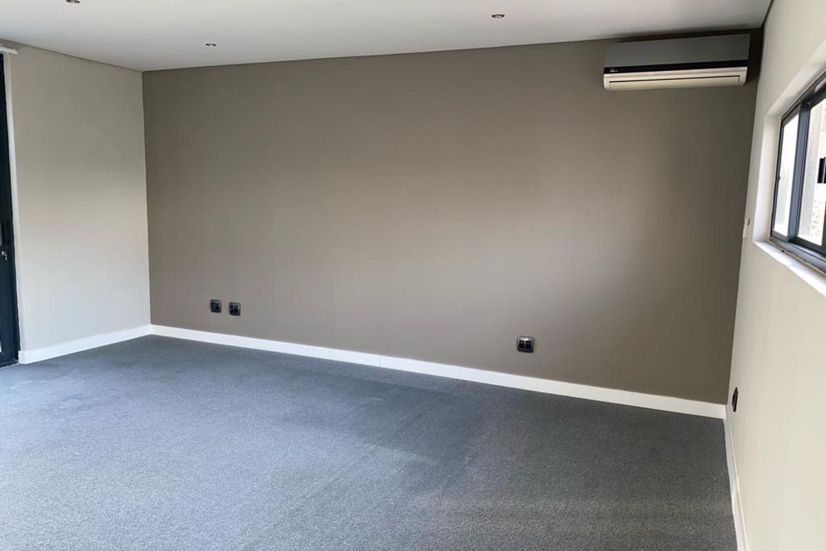 A clean, empty bedroom featuring neutral wall tones, grey carpeting, recessed lighting, and a wall-mounted split-system air conditioning unit.